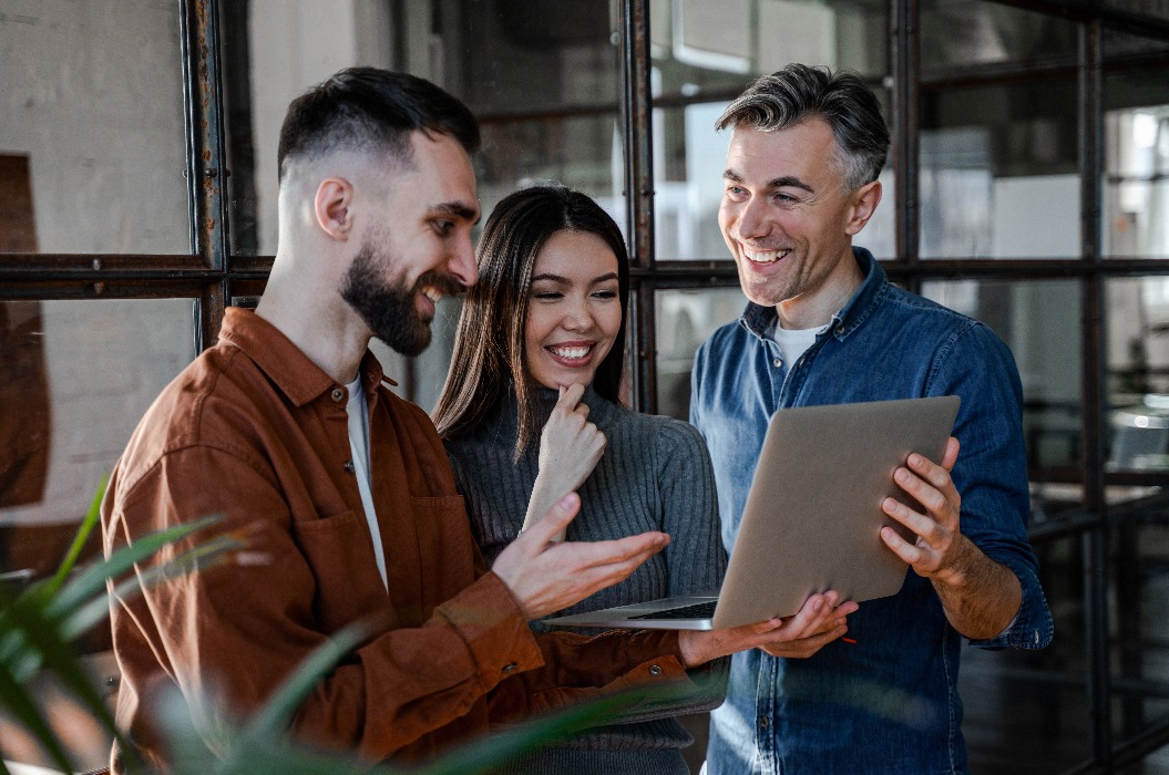 Drei glückliche Kollegen arbeiten am Laptop und diskutieren, Teamarbeit, kreativer Prozess. Erfolgreiches Konzept für eine Geschäftsstrategie. Selbstbewusster Büroangestellter in einem Coworking Space.